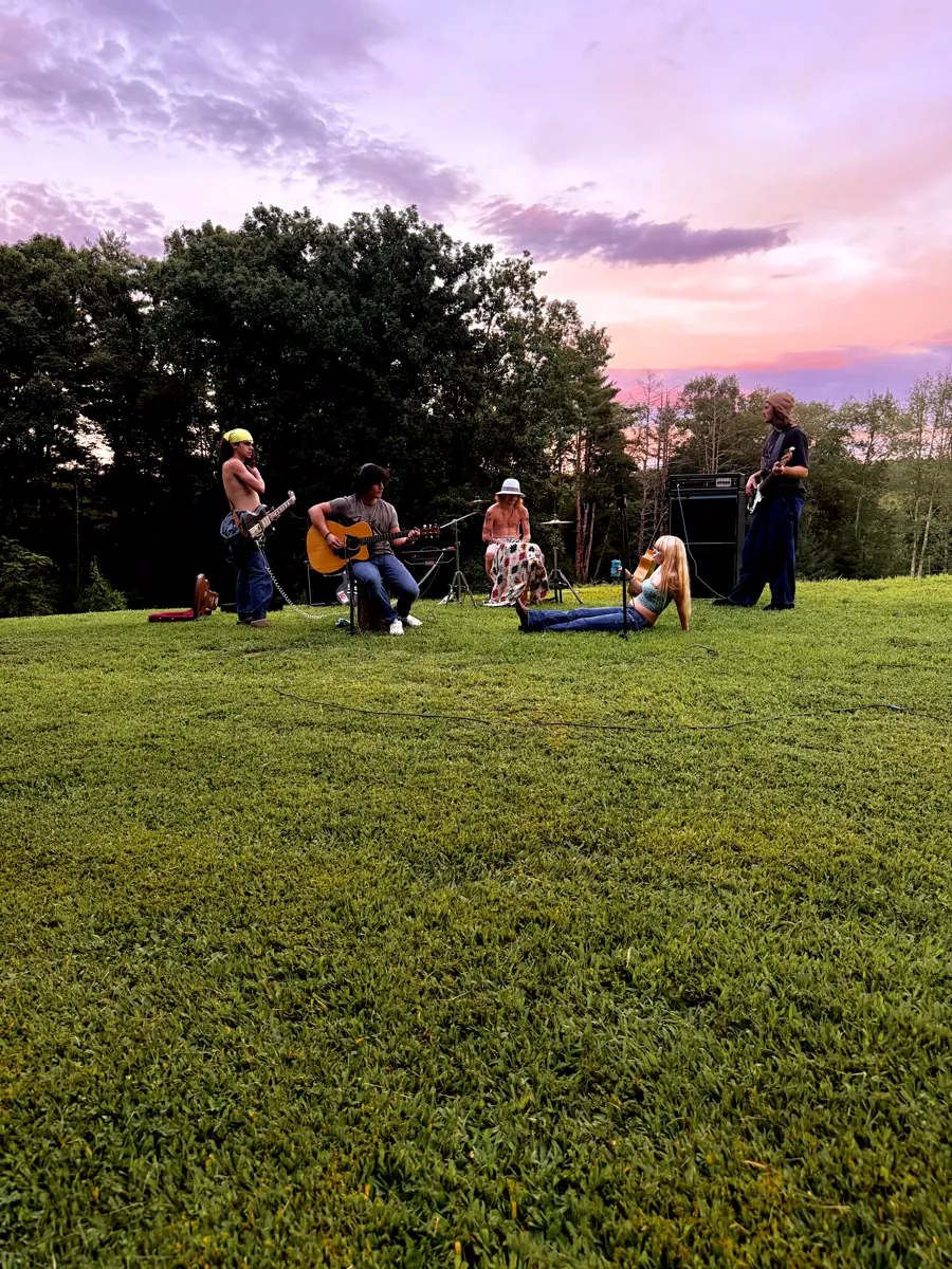 The band set up outdoors on a mown hilltop at dusk, two guitarists and a drummer on the grass with the singer sitting out front and a pink and violet sunset behind the treeline
